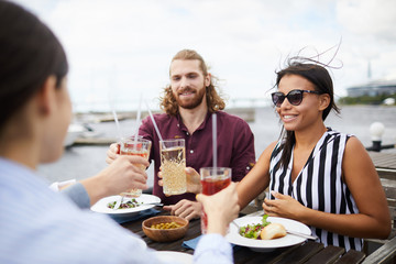 Young restful men and women cheering up by drinks while sitting by table during lunch