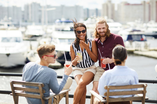 Young joyous couple with drinks sitting by waterside, having talk and relaxing