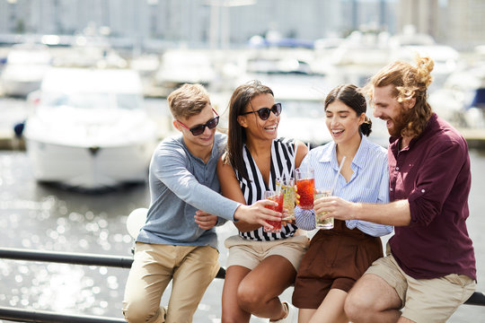 Laughing girls and their boyfriends clinking with glasses of drinks while sitting by waterside on summer day