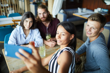 Group of four young friends making selfie while sitting by table and relaxing in cafe