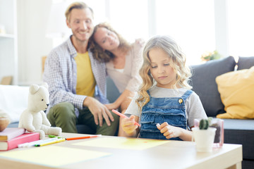 Cute little girl with curly hair drawing with highlighters with her parents on background