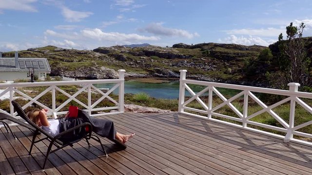 A woman reading a book lying in a trestle on a wooden terrace with a view of the Norwegian fjord in sunny weather, Averoy, Norway
