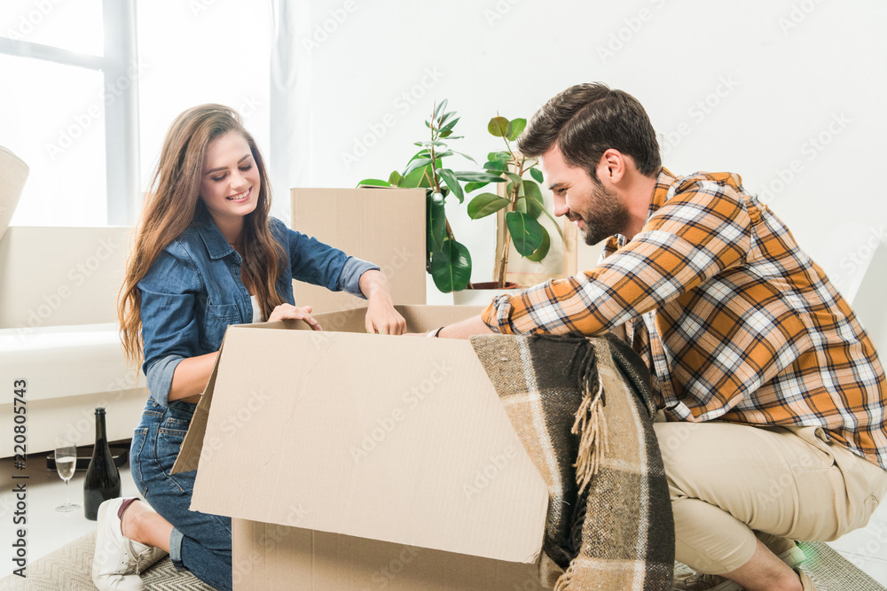 Wall mural smiling couple unpacking cardboard boxes together at new home, moving home concept