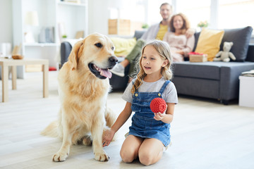 Little girl holding red rubber toy while sitting next to labrador pet and playing with him
