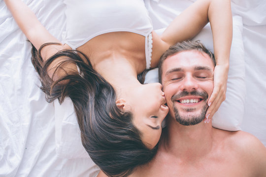 Gorgeous and attractive couple is lying in bed face to face. Guy keeps his eyes closed and smiles. Girl holds her hand on his cheek and kisses him.