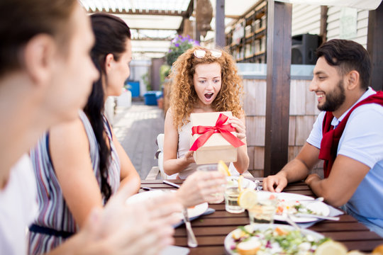 Portrait of group of friends celebrating birthday in outdoor cafe, focus on excited young woman opening gift box with red ribbon, copy space