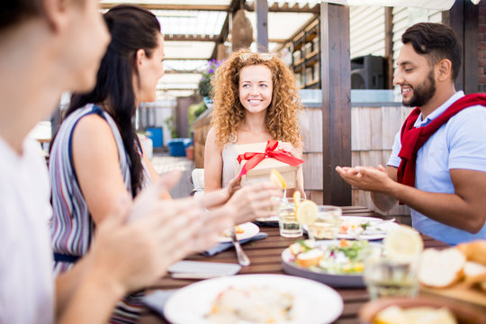 Portrait of group of friends celebrating birthday in outdoor cafe, focus on happy young woman holding gift box with red ribbon, copy space
