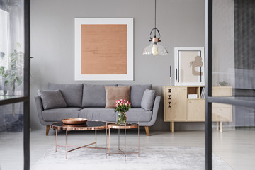 Flowers on copper table in front of grey couch in living room interior with rose gold poster. Real photo