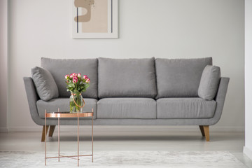 Flowers on copper table in front of grey sofa in bright living room interior with poster. Real photo
