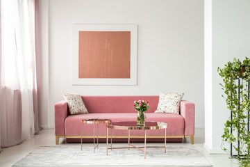 Pillows on pink sofa in white apartment interior with painting and flowers on copper table. Real...
