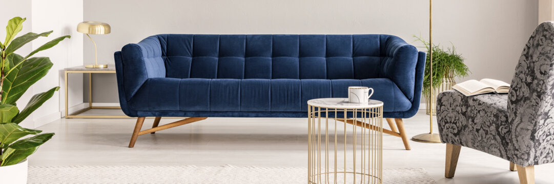 Real photo of a dark blue sofa. golden coffee table, plant and patterned armchair with a book in a living room interior