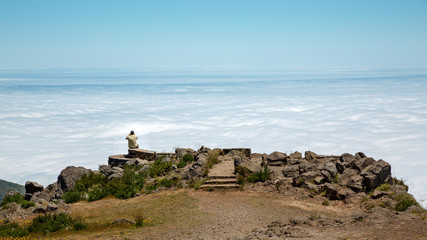 Man sitting with his back on edge of cliff above the clouds and takes photo.