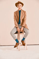 Young male model with red hair and freckles posing in brown coat and hat, plaid pants, turquoise...