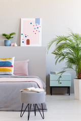 Abstract poster in real photo of bright bedroom interior with open book on hairpin pouf, fresh plants and bedside table