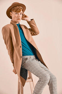 Young male model with red hair and freckles in brown coat and hat, plaid pants, turquoise sweater and socks, sitting on chair. Creamy background