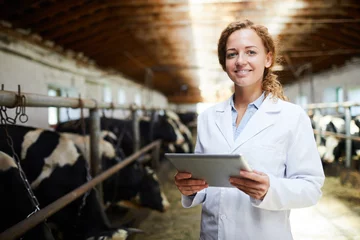 Foto auf Acrylglas Tierärzte Taillenporträt einer fröhlichen Tierärztin, die lächelt und in die Kamera schaut, während sie ein digitales Tablet verwendet, das im Kuhstall eines modernen Milchviehbetriebes steht, Kopierraum