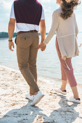 partial view of romantic couple holding hands while walking on sandy riverside