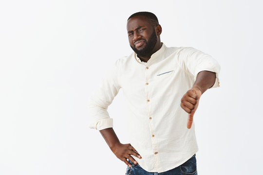 This place sucks, giving my dislike. Portrait of unimpressed displeased gloomy dark-skinned man in white shirt, grimacing, wrinkling nose and showing thumbs down from digust and negative opinion