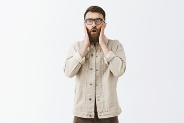 Studio shot of emotive shocked and worried male model with long beard in glasses holding hands on face looking nervous and shook learning terrifying bad news expressing empathy while in stupor