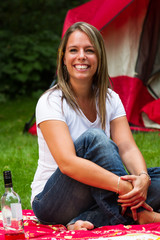 Smiling Beautiful Young Woman Sitting on a Blanket on the Grass, with a Tent Behind Her and a Wine Bottle on the Blanket