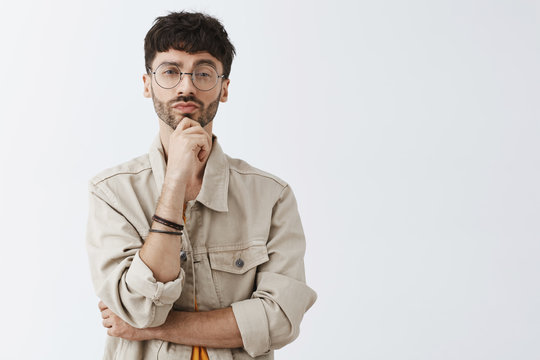 Intrigued stategist liking your suggestion. Smart and creative thoughtful male entrepreneur in glasses and beige jacket standing in hmm pose with hand on chin and folded lips thinking making decision