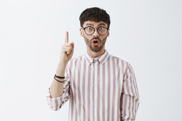 Waist-up shot of thrilled and excited attractive young hipster guy in glasses and pink shirt raising index finger in eureka finger folding lips, gasping adding important suggestion having great idea