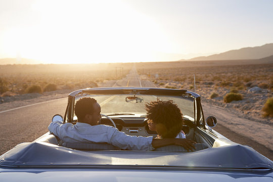 Rear View Of Couple On Road Trip Driving Classic Convertible Car Towards Sunset