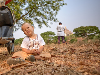 Cute boy in local burmese clothes resting sitting on the dry foliage by the e-bike while his mother takes pictires of the Thatbyinnyu Temple in Bagan, Myanmar