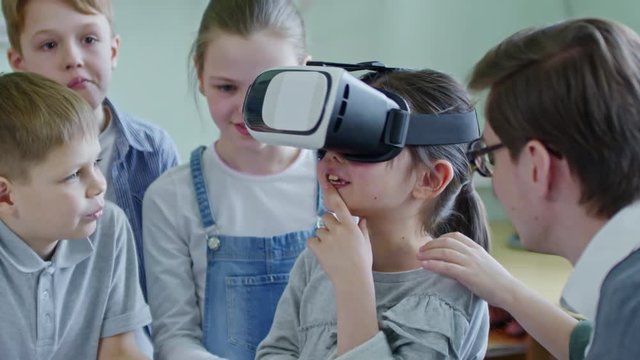 Medium shot of cute girl of primary school age using virtual reality goggles, her classmates standing around and looking at her