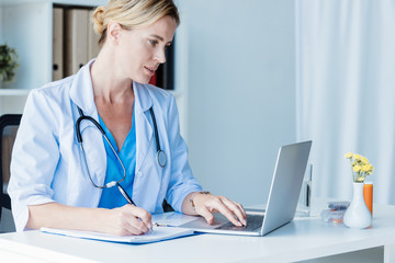 focused female doctor writing in clipboard at table with laptop in office