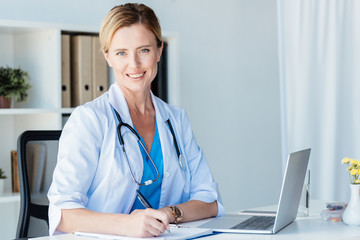 smiling female doctor looking at camera while writing in clipboard at table with laptop in office