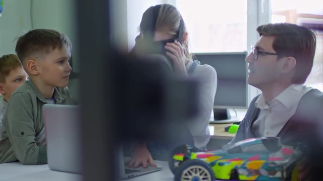 Medium shot of young girl of elementary school age exploring virtual worlds with vr headset during information technology lesson at school, teacher and classmates looking at her