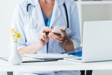 cropped image of female doctor using smartphone at table with laptop in office