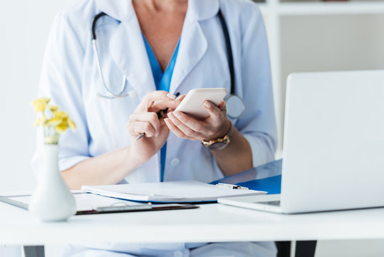 cropped image of female doctor using smartphone at table with laptop in office