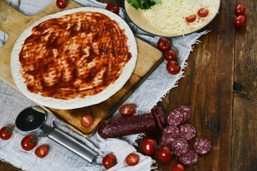 Ingredients for Pizza: basis dough, salami sausage, cherry tomatoes, grated cheese, tomato paste, herbs and arugula. Natural Wooden Background With Linen Napkin and Knife For Pizza