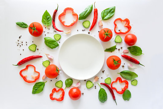 Colorful salad ingredients pattern made of tomatoes, pepper, chilli, garlic, cucumber slices, basil and empty plate on white background. Cooking concept. Top view. Flat lay. Copy space