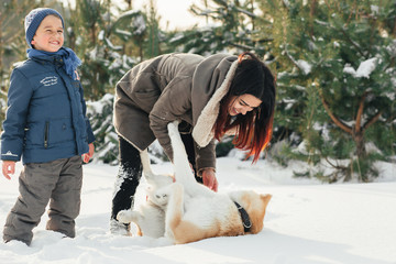 Little child and mom plays with funny Akita-inu dog in a winter park. Christmas happy family,mother and son walking with dog lying on snow in winter day. Drinking hot coffee or tea on snowy winter