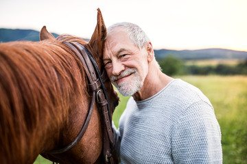 A senior man standing close to a horse outdoors in nature, holding it.