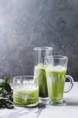 Matcha green tea iced latte or cocktail in three different glasses with ice cubes, matcha powder and jug of milk on white marble table, decorated by green branches. Grey wall at background