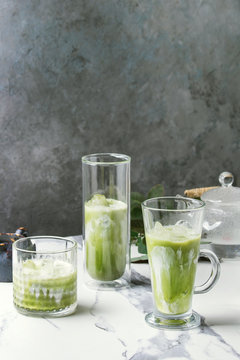 Matcha green tea iced latte or cocktail in three different glasses with ice cubes, matcha powder and jug of milk on white marble table, decorated by green branches. Grey wall at background