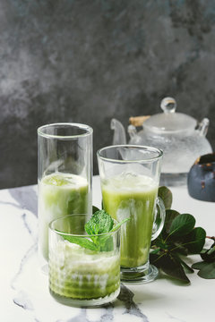 Matcha green tea iced latte or cocktail in three different glasses with ice cubes, matcha powder and jug of milk on white marble table, decorated by green branches. Grey wall at background