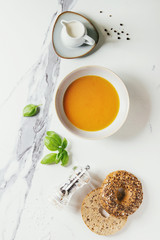 Bowl of vegetarian pumpkin carrot soup served with herbs, spoon, jug of cream, bagel bread, black pepper, salt over white marble texture background. Flat lay, space