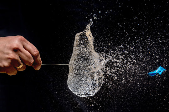 Bursting Of A Blue Balloon Filled With Water With A Long Pin On Black Background. 