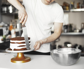 Close up of young man smearing cream on chocolate cake by metal spatula in hand.