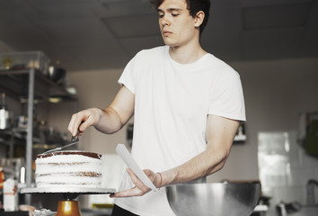Close up of young man smearing cream on chocolate cake by metal spatula.