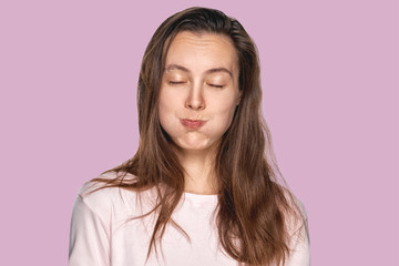 Funny pretty girl puffing out her cheeks against lavender studio background. Headshot of charming red haired young woman making mouths while having fun indoors. People, lifestyle, youth and happiness
