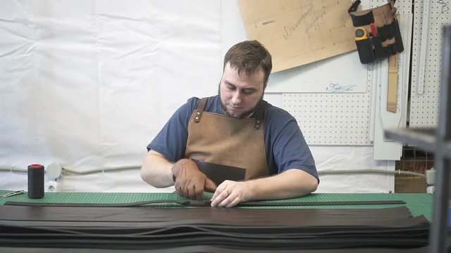 Male leather worker sewing black long detail in studio. Craftsman working in brown dickey and glove on his right hand, where he holding the needle. Portrait. Indoors.