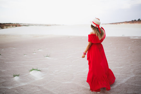 Beautiful young girl in red dress and fedora hat with stripe walking in the desert with white sand