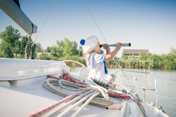 Obraz premium Funny little baby captain on board of sailing yacht watching offshore sea on summer cruise. Travel adventure, yachting with child on family vacation. Kid clothing in sailor style, nautical fashion.