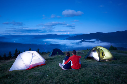 Back view, loving people sitting near camping enjoying sunrise in the mountains. In the valley between the mountains the lights of the town are burning which are covered with morning mountain haze